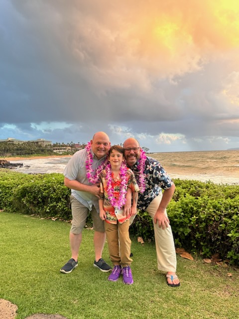 Two dads and a child and leis on a beach
