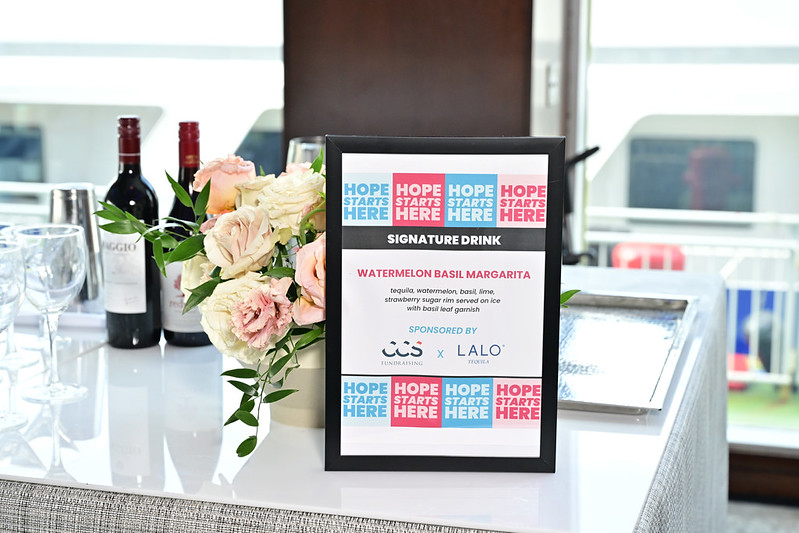 Sign advertising a signature drink sponsor on a table with wine bottles and roses.